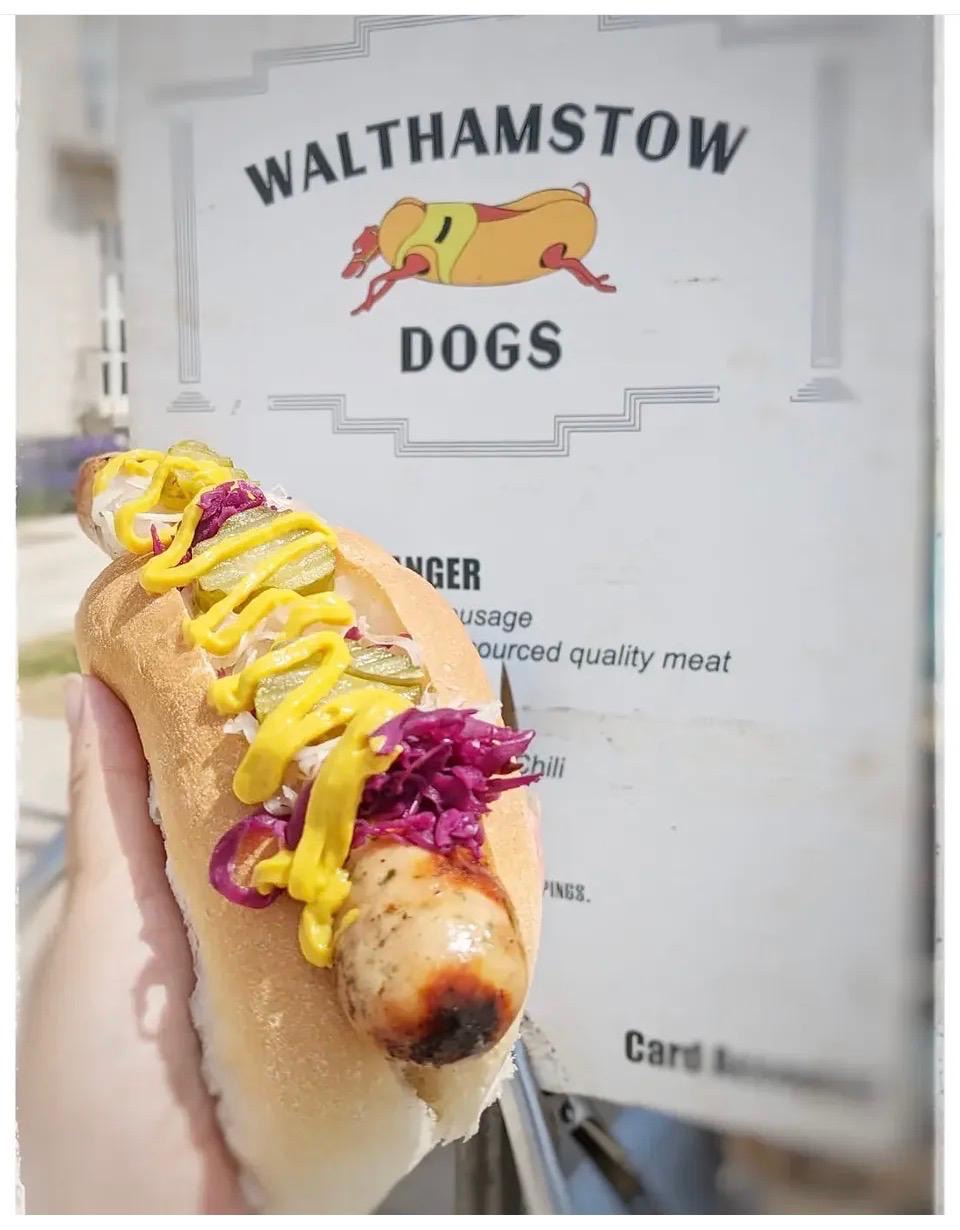 Hot dog loaded with pickles, mustard and red cabbage held in front of the Walthamstow Dogs menu